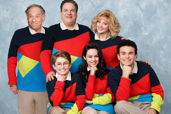 Photo of the Goldbergs. They are all in the same matching geometric bold coloured sweater with the adults standing and the kids sitting on a bench not visible to the camera. The kids have their right fist under their chins with their right leg crossed over their other leg.