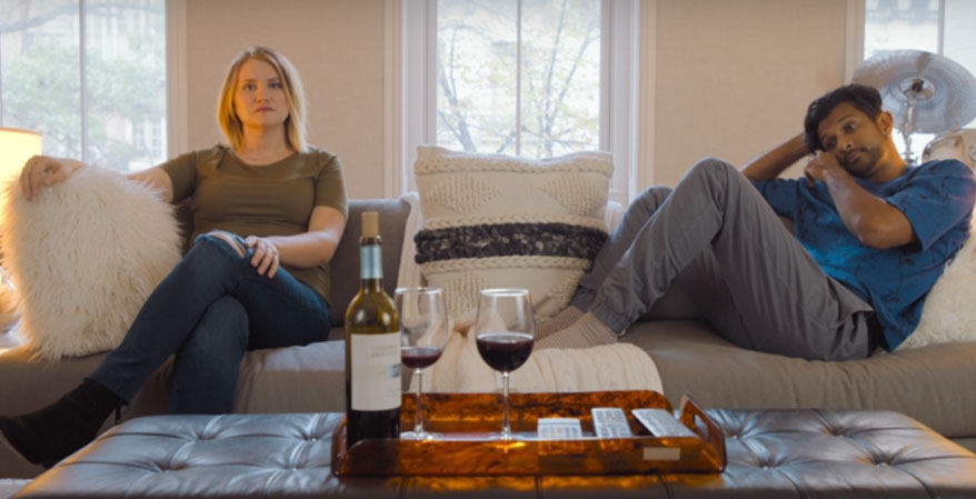 Brittany sitting on the couch with Jern. Two adults drinking wine on a couch. Woman sitting upright, fully dressed with legs crossed. Man with legs and feet on the couch slouching on the couch.