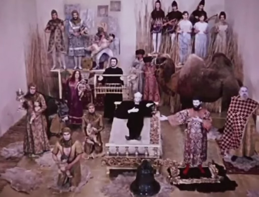 Screenshot of poet's dream. From the back: knights on a table, a chorus and lute players, man holding a swan, large woman holding a baby in purple, man holding a cage, man holding robe to a camel. From the front left: a calf, a woman in beige holding a candle and a baby, two woman knelling holding a baby. From the right: man standing on rug with plaid shroud, man with right arm extended outwards and left arm on his chest standing on a rug. Centre: Woman in black with a crown laying down extending her left arm on a bed in front of a bell.