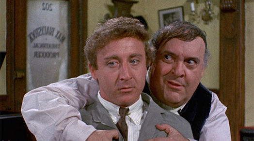 Screen shot of The Producers. Max Bialystock is conning Leopold Bloom in his office. Max is partially hugging Leo with crazed look in his eyes. Leo looks sad and looking to the bottom left corner, not at Max. Both are facing the camera.