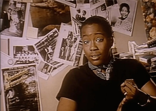 Cheryl is talking to her camera in front of a wall with photos of the unknown actress sitting down.