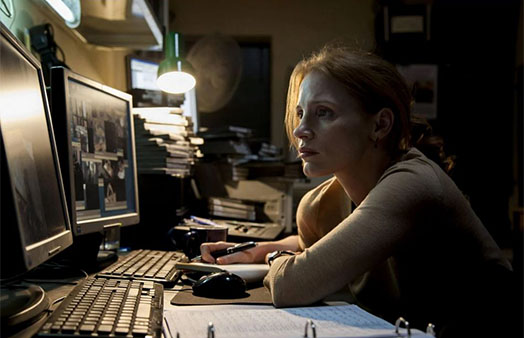 Woman sitting at a computer desk with two monitors and two keyboards with multiple video tapes in the background.